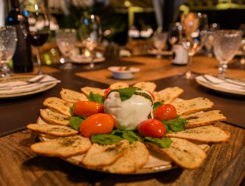 Foto profissional de uma Burrata rodeada de tomates pequenos, sobre folhas frescas de rúcula e com torradas douradas em volta. Sobre a Burrata, há folhas de Manjericão para finalizar a decoração do prato.O prato está montado em uma mesa de madeira com taças de vinho e pratos desfocados ao fundo. O ambiente está decorado para um evento no restaurante Botegare, em Penedo, RJ. Penedo é um roteiro conhecido por sua culinária no aluguel por temporada.