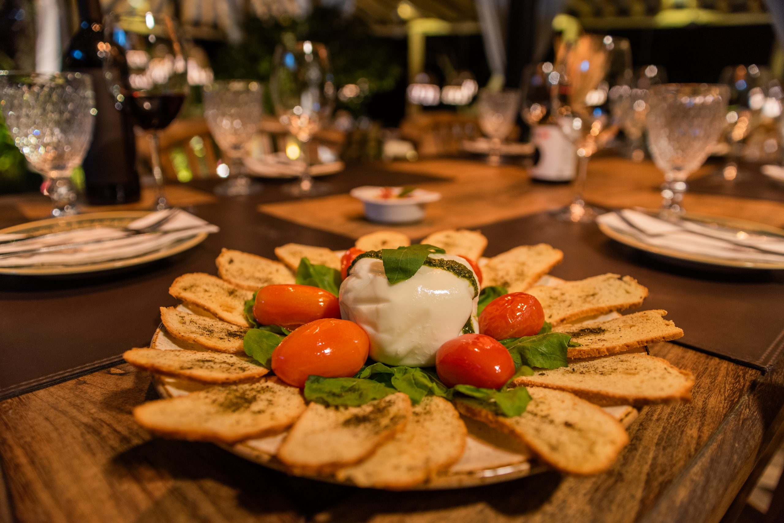 Foto profissional de uma Burrata rodeada de tomates pequenos, sobre folhas frescas de rúcula e com torradas douradas em volta. Sobre a Burrata, há folhas de Manjericão para finalizar a decoração do prato.O prato está montado em uma mesa de madeira com taças de vinho e pratos desfocados ao fundo. O ambiente está decorado para um evento no restaurante Botegare, em Penedo, RJ. Penedo é um roteiro conhecido por sua culinária no aluguel por temporada.