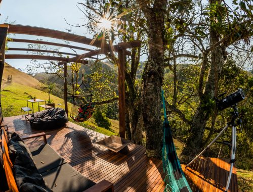 Foto profissional de um deck de madeira na parte externa de um chalé-container instalado no alto de uma montanha de Visconde de Mauá, RJ. O deck tem uma rede para duas pessoas, um sofá de madeira grande com alfmofadas pretas, um puff grande e preto. Tem também um pergolado coberto com vidro, onde uma cadeira de balançar de tecido colorida está pendurada, ao lado de uma mesa e dois bancos de metal e madeira. Uma árvore faz sombra no deck, e ao fundo, vê-se outras montanhas da região. O ambiente foi preparado para receber pessoas que fazem home office, unindo viagem e trabalho.