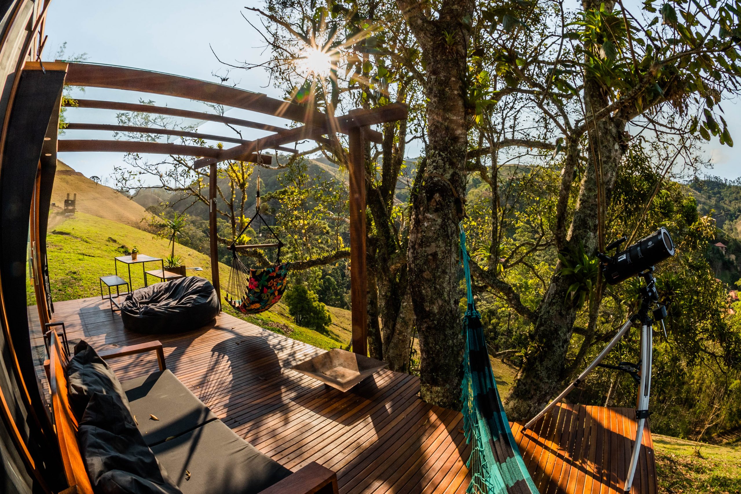 Foto profissional de um deck de madeira na parte externa de um chalé-container instalado no alto de uma montanha de Visconde de Mauá, RJ. O deck tem uma rede para duas pessoas, um sofá de madeira grande com alfmofadas pretas, um puff grande e preto. Tem também um pergolado coberto com vidro, onde uma cadeira de balançar de tecido colorida está pendurada, ao lado de uma mesa e dois bancos de metal e madeira. Uma árvore faz sombra no deck, e ao fundo, vê-se outras montanhas da região. O ambiente foi preparado para receber pessoas que fazem home office, unindo viagem e trabalho.